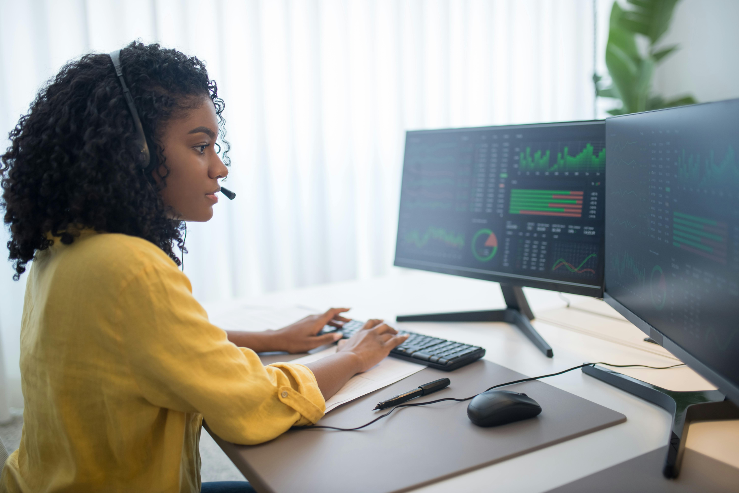 African American woman working in office analyzing data on dual computer monitors.